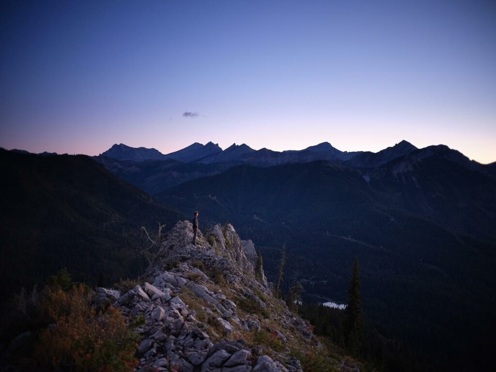 A man standing on a craggy peak at sundown. High, snow-capped mountain peaks in the distance.