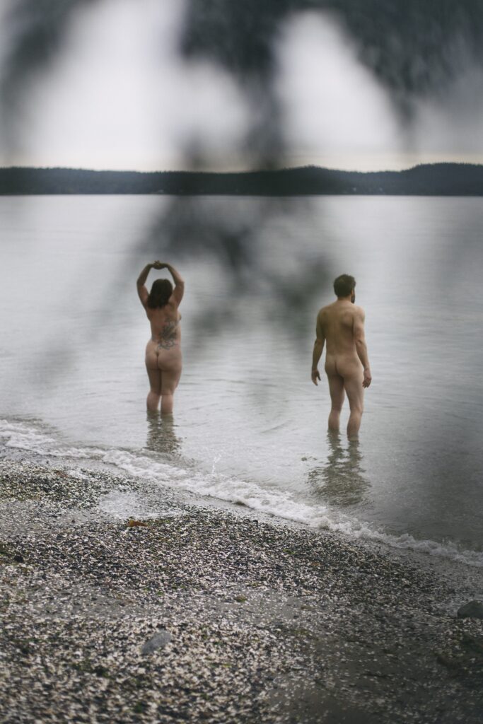 Two naked mature white people standing in sea surf with pebble beach behind them