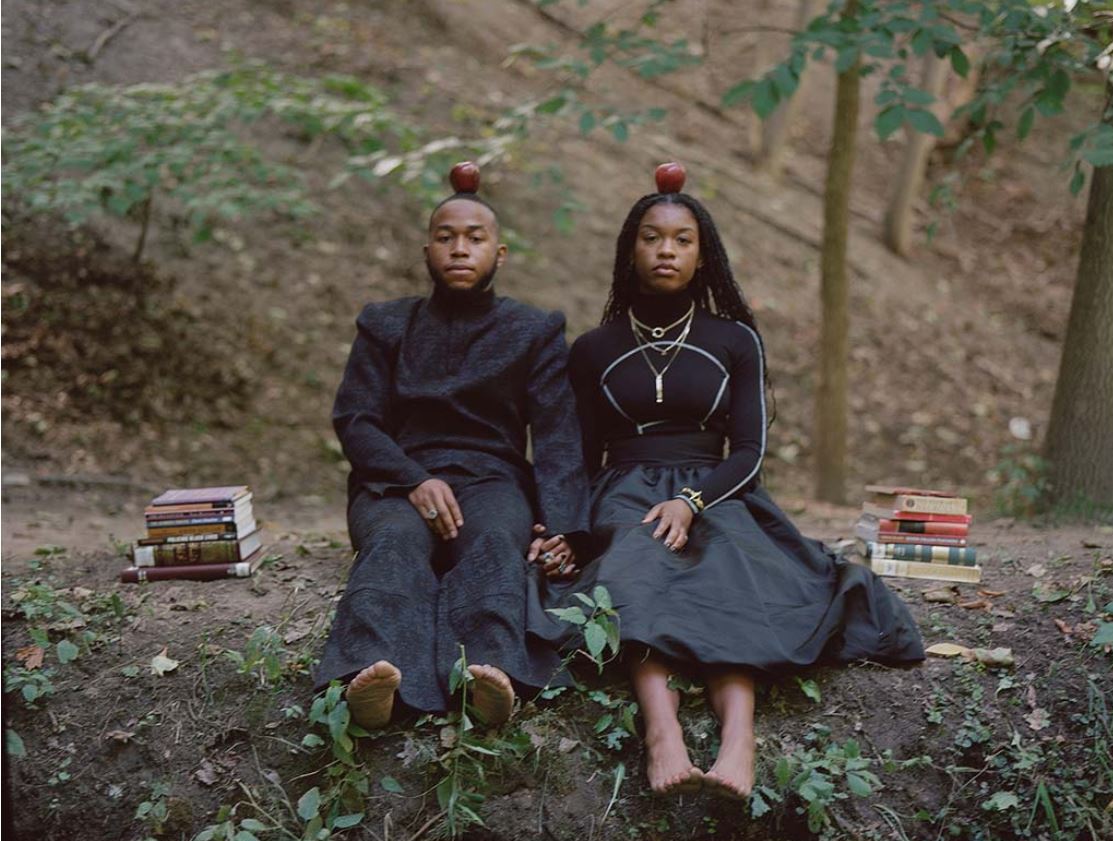 Black couple sitting on the ground outside with apples on their head, holding hands, stacks of books on both sides