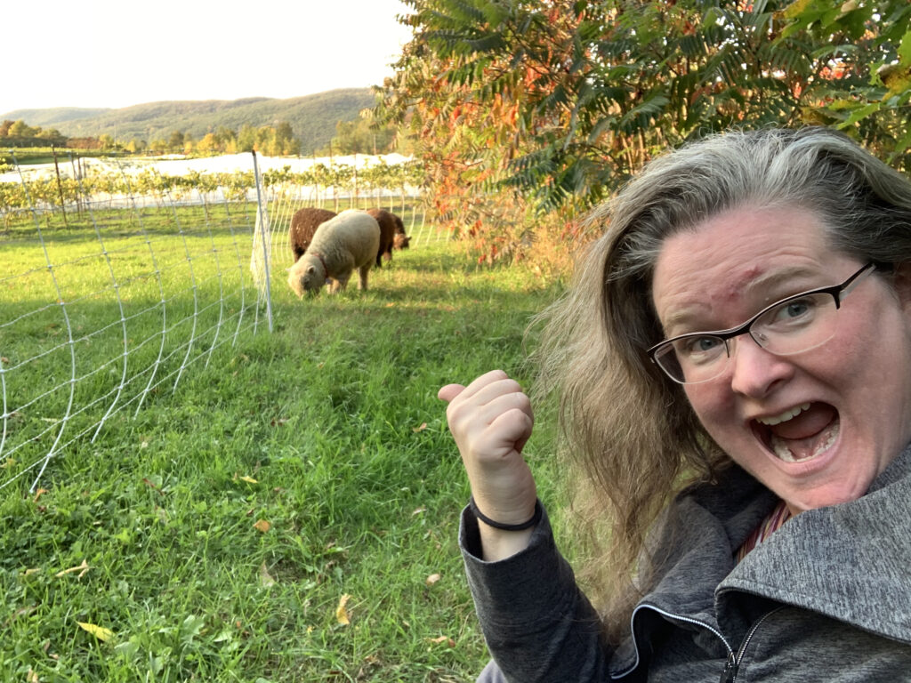 Woman in right foreground, mouth open, pointing her right thumb back at the pasture behind her with a couple of sheep grazing and a tree in early fall colours