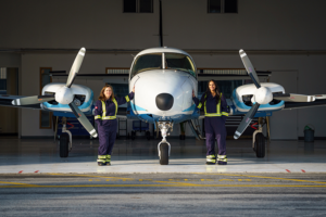 Frontal view of two women pilots in coveralls standing beside a twin prop plane, each with a hand on the fuselage.