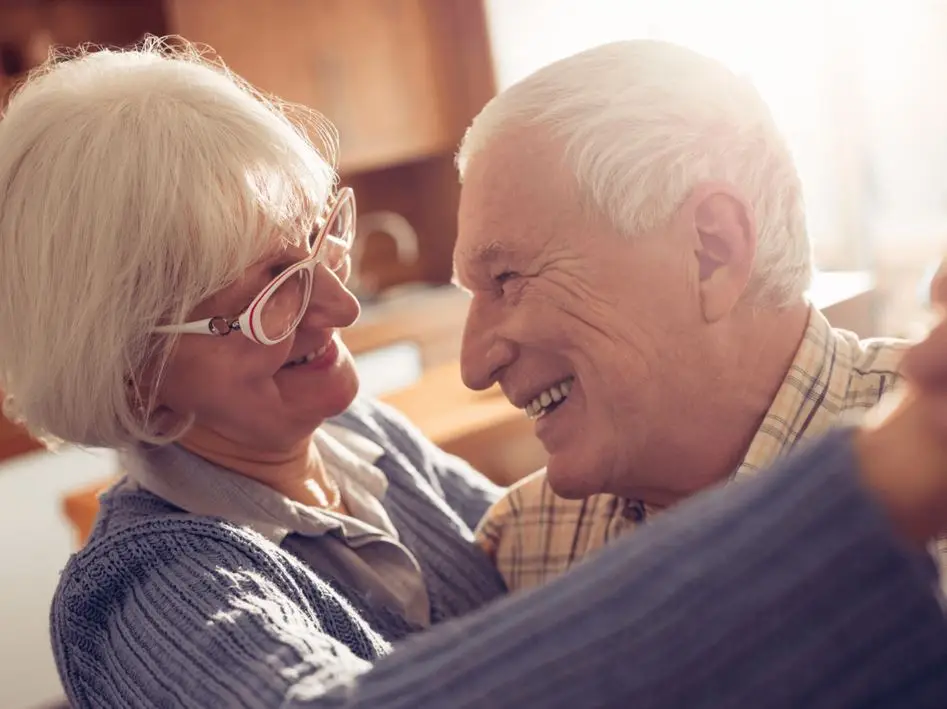 An elderly couple smiling at each other while dancing in their kitchen