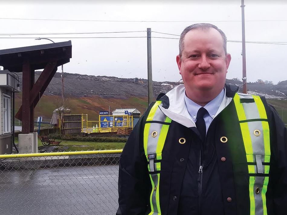 Portrait of Ben Routledge in a safety vest at right front of image with light industrial landscape behind him