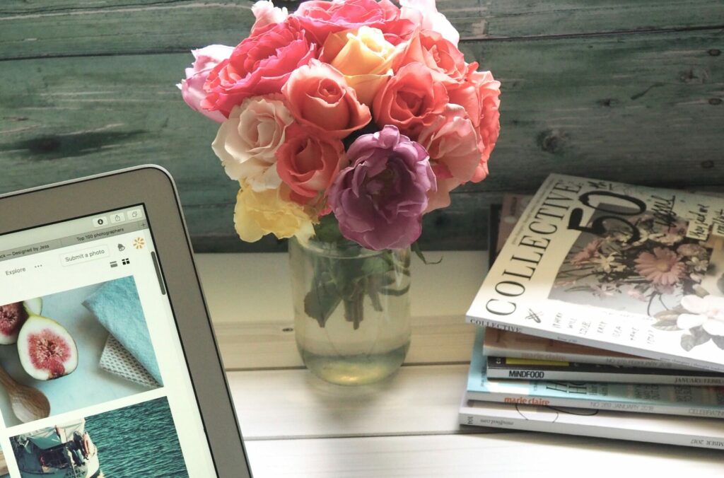 Pile of magazines, digital clock, laptop with images, clear vase with flowers.