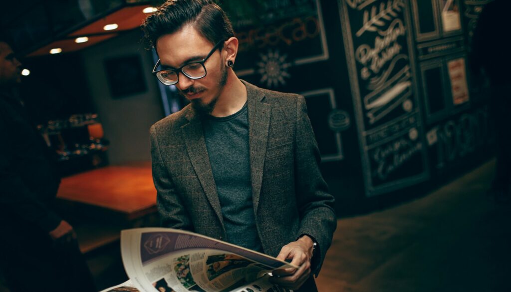 Man wearing blazer paging through publication. Bar or lobby in background. Photo by Helena Lopes: https://www.pexels.com/photo/man-wearing-a-blazer-2017803/