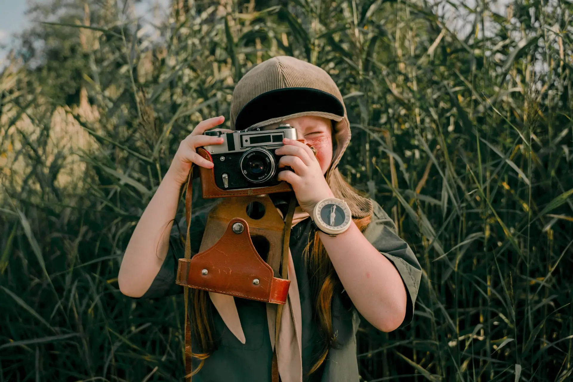 Girl with hat holding old camera with leather case below pointed toward viewer with tall grasses behind