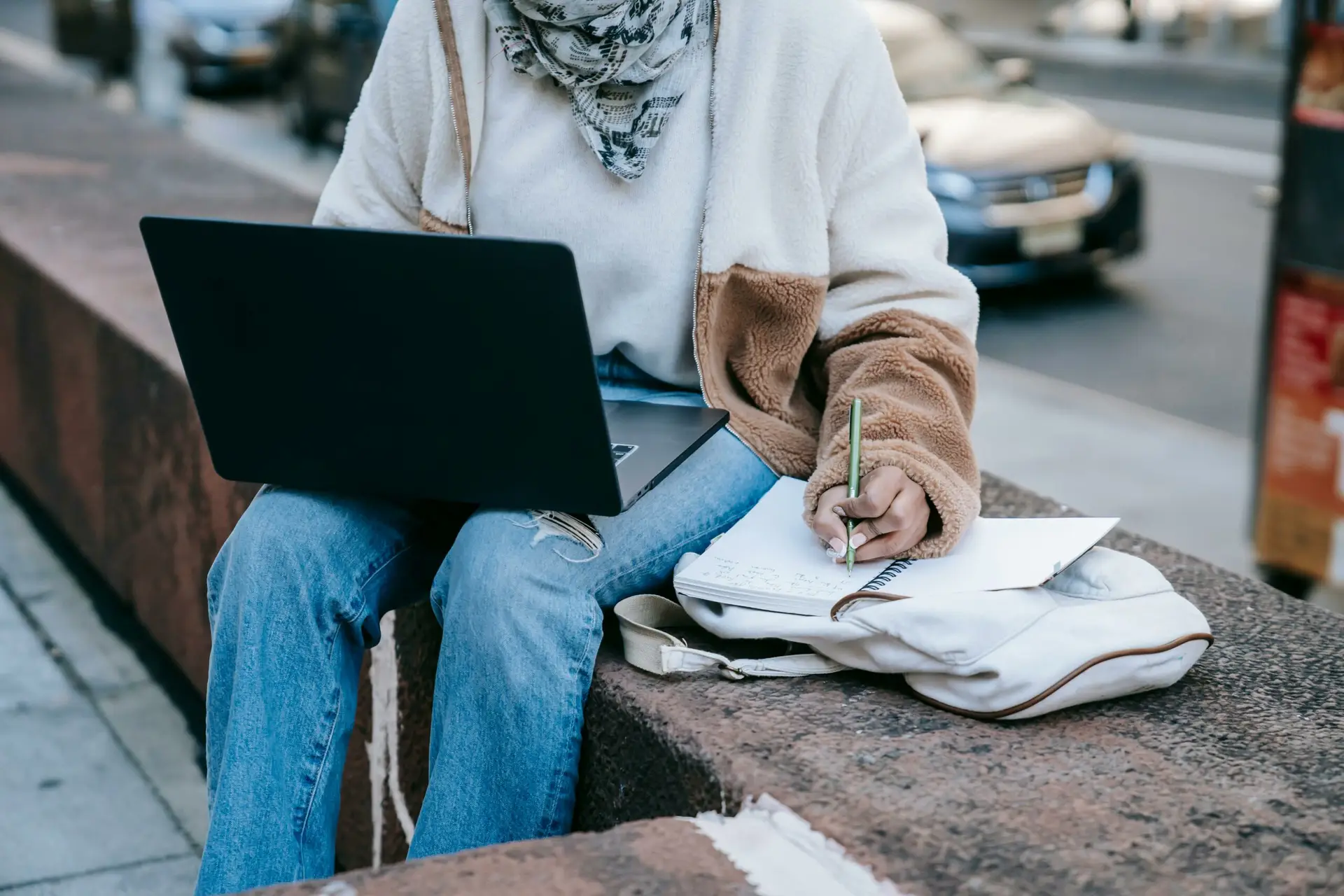 Person wearing bluejeans and a sweater sitting outside on concrete ledge making notes on pile of papers with laptop balanced on lap.