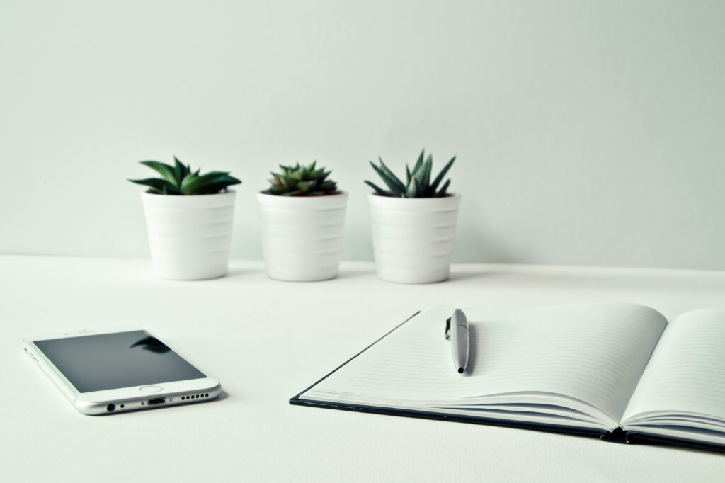 On a white desktop 3 small white planters in background with succulents, a white cellphone on the left and a large, open notebook with a pen on the right.