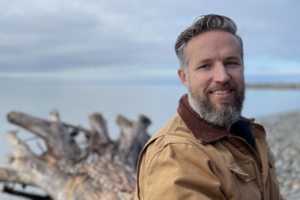 Anthony Robinson on beach in front of tree stump