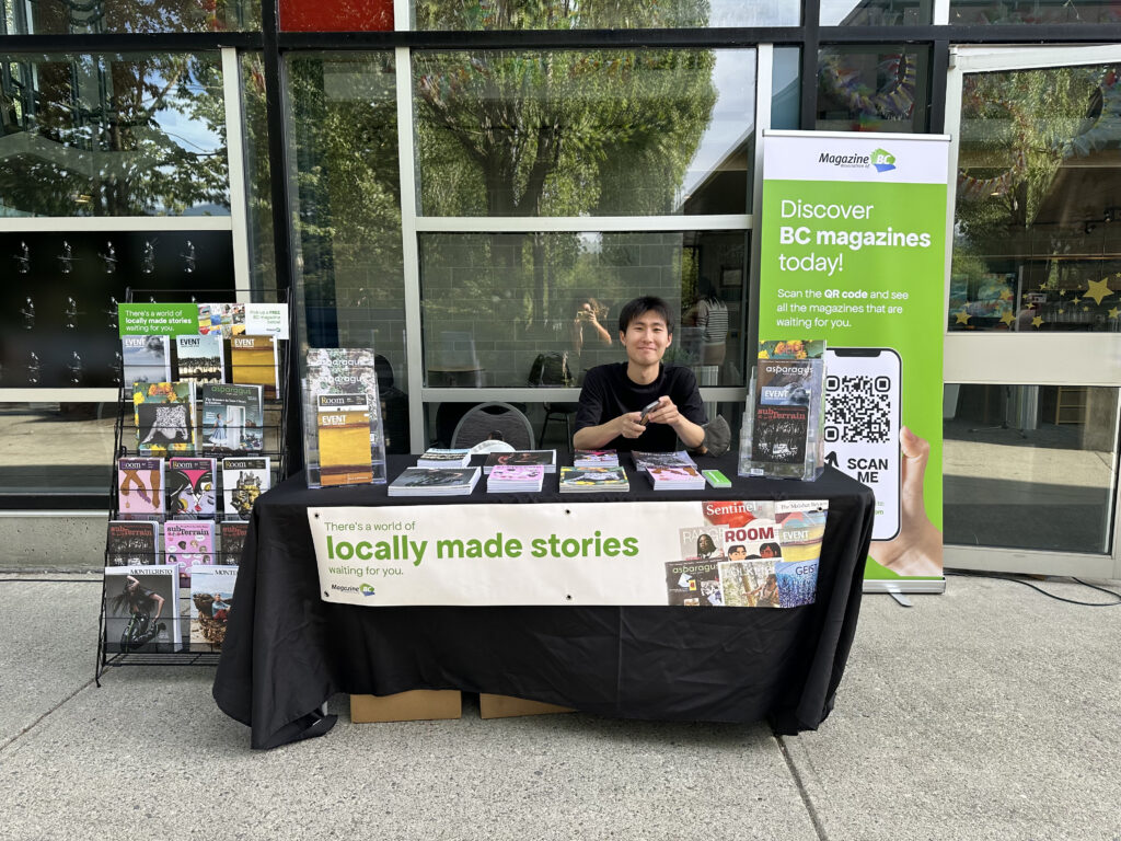 Sun Woo Baik sitting at a table at Evergreen Cultural Centre Coquitlam displaying MagsBC member magazines including a rack of magazines to his right and a banner to his left, reading: "Discover BC magazines today. Scan the QR code and see all the magazines that are waiting for you. Also, at front of table: There's a world of locally made stories waiting for you.