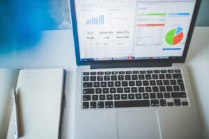 Image by Pixabay showing silver laptop open with screen of graphs and pie chart, open notebook with pen to the left