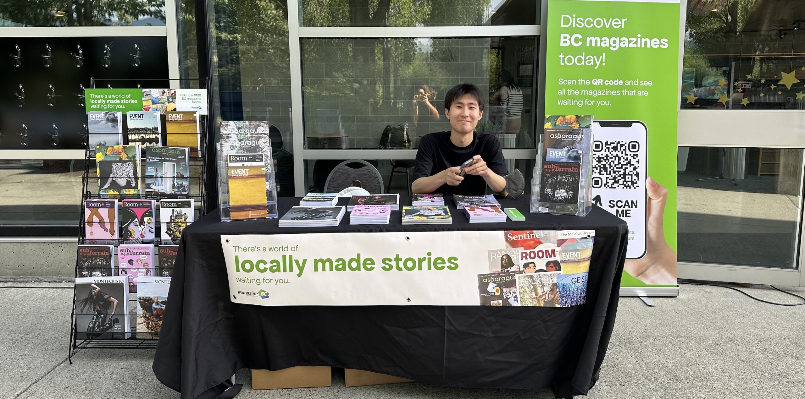 Sun Woo Baik sitting at a table at Evergreen Cultural Centre Coquitlam displaying MagsBC member magazines including a rack of magazines to his right and a banner to his left, reading: "Discover BC magazines today. Scan the QR code and see all the magazines that are waiting for you. Also, at front of table: There's a world of locally made stories waiting for you.