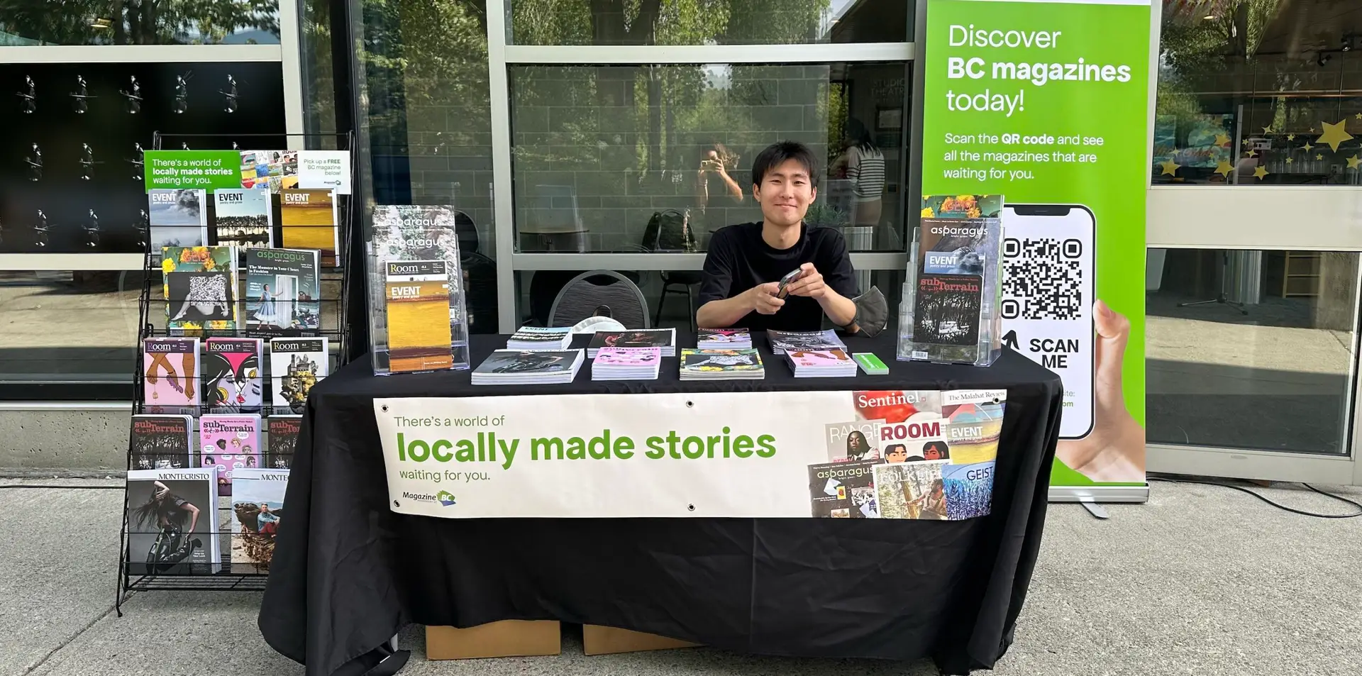 Sun Woo Baik sitting at a table at Evergreen Cultural Centre Coquitlam displaying MagsBC member magazines including a rack of magazines to his right and a banner to his left, reading: "Discover BC magazines today. Scan the QR code and see all the magazines that are waiting for you. Also, at front of table: There's a world of locally made stories waiting for you.