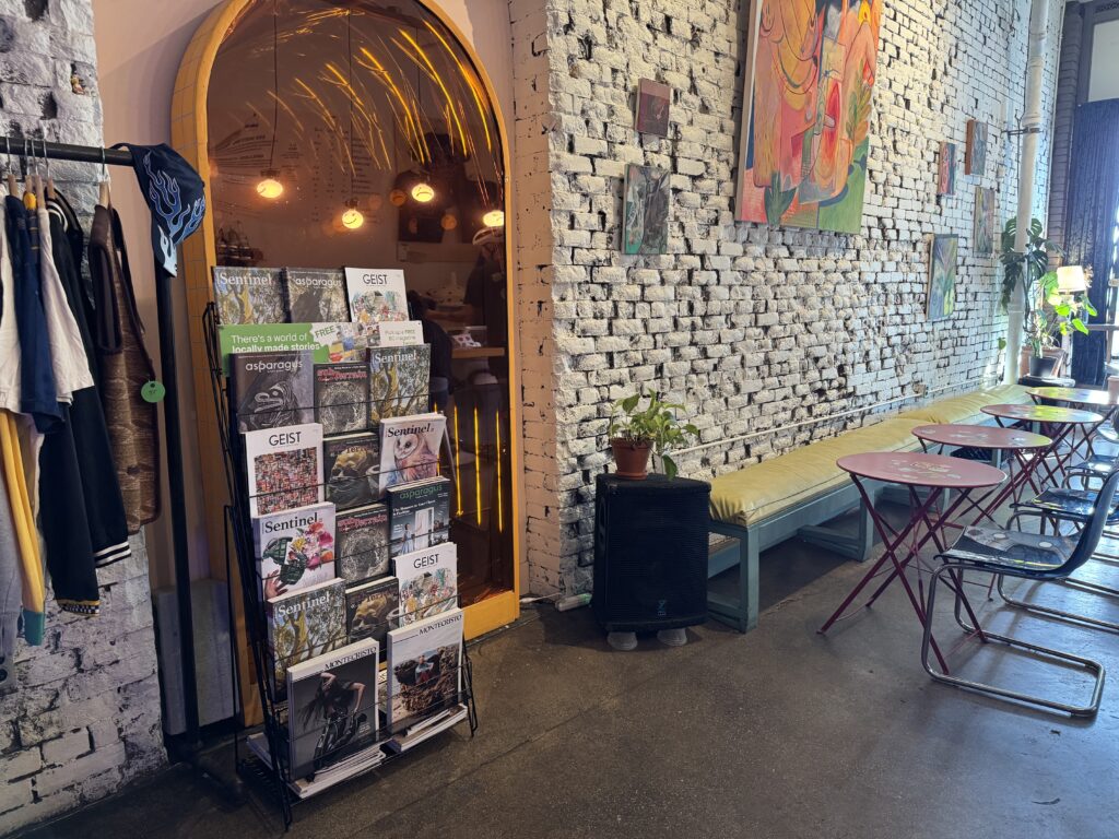 Featuring a display rack of BC magazines at Moodswing Coffee + Bar, New Westminster, as well as a coat rack on the left and empty seating on the right with various paintings on the brick wall above.
