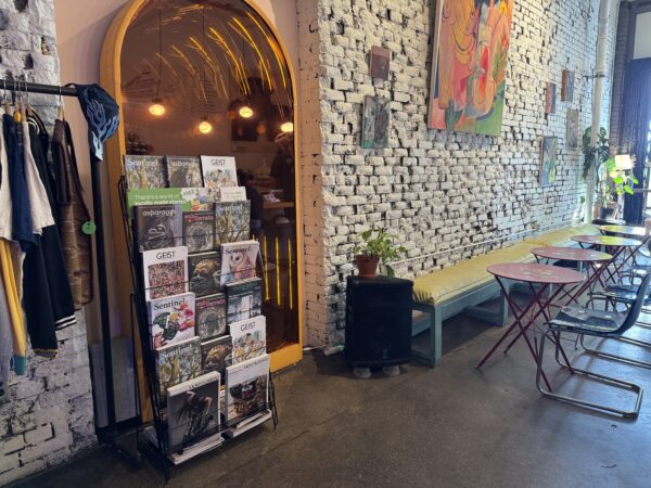 Featuring a display rack of BC magazines at Moodswing Coffee + Bar, New Westminster, as well as a coat rack on the left and empty seating on the right with various paintings on the brick wall above.