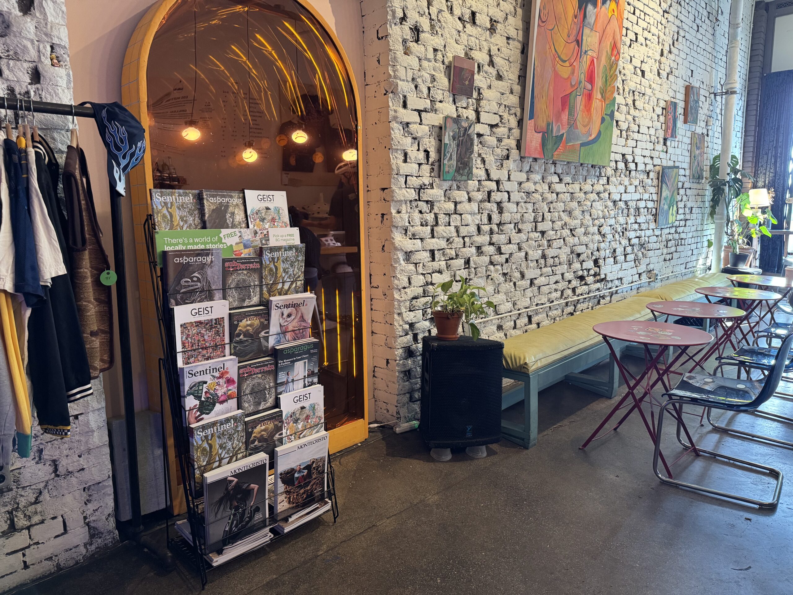 Featuring a display rack of BC magazines at Moodswing Coffee + Bar, New Westminster, as well as a coat rack on the left and empty seating on the right with various paintings on the brick wall above.