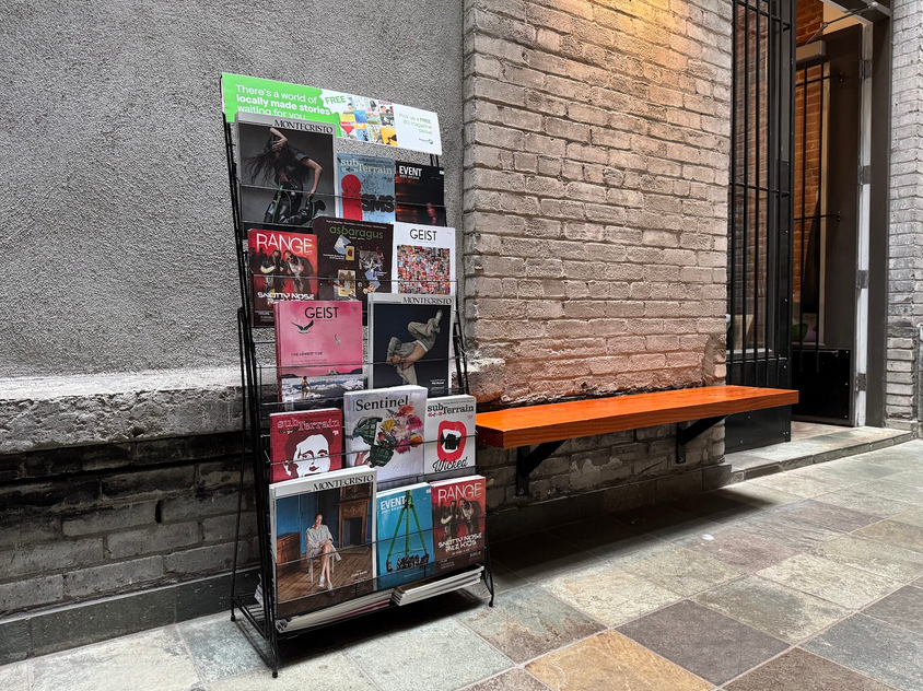 Display rack of BC magazines against a cement and brick wall at Saunter Coffee, downtown Vancouver, with an open door to the right.