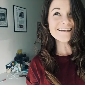 Colour portrait of Shelby Blackley, smiling. In the background what appears to be a desk with several items and pictures on the wall above.