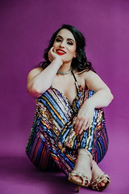 A full-size colour portrait of Jessica Key, seated on the ground on a magenta, seamless background facing forward and smiling into the camera. She is cupping her face with her right arm and wears a colourful, glittering outfit.