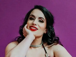 A full-size colour portrait of Jessica Key, seated on the ground on a magenta, seamless background facing forward and smiling into the camera. She is cupping her face with her right arm and wears a colourful, glittering outfit, tight crop